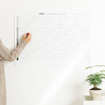 A persons hand writing on the A1 2025 A Lot Can Happen In A Year Wall Planner, the planner is hanging on the wall and there is a plant on the table next to it.