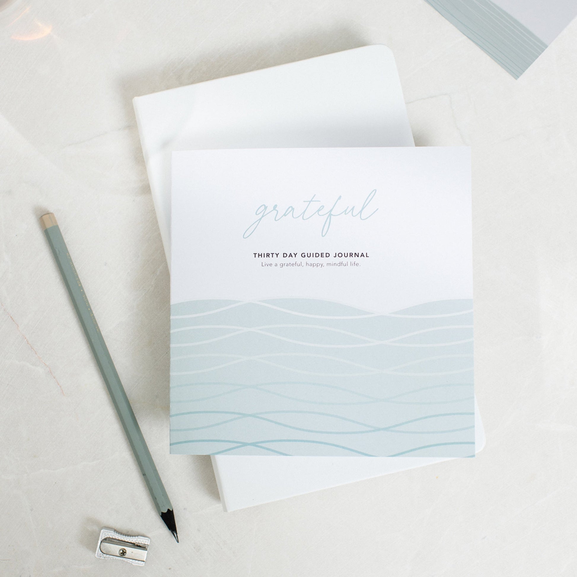 Grateful guided gratitude journal, placed on top of another journal with a pencil and pencil sharpener next to it