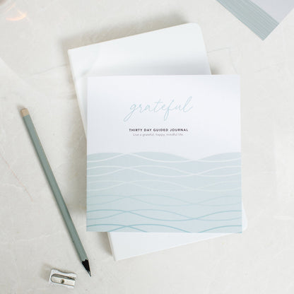 Grateful guided gratitude journal, placed on top of another journal with a pencil and pencil sharpener next to it