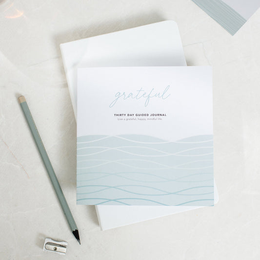Grateful guided gratitude journal, placed on top of another journal with a pencil and pencil sharpener next to it