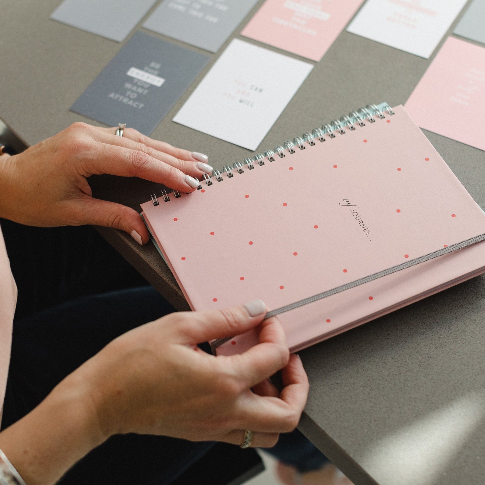 A woman's hand holding the pink ivf journal it has a spot pattern on the cover and the words IVF Journey...