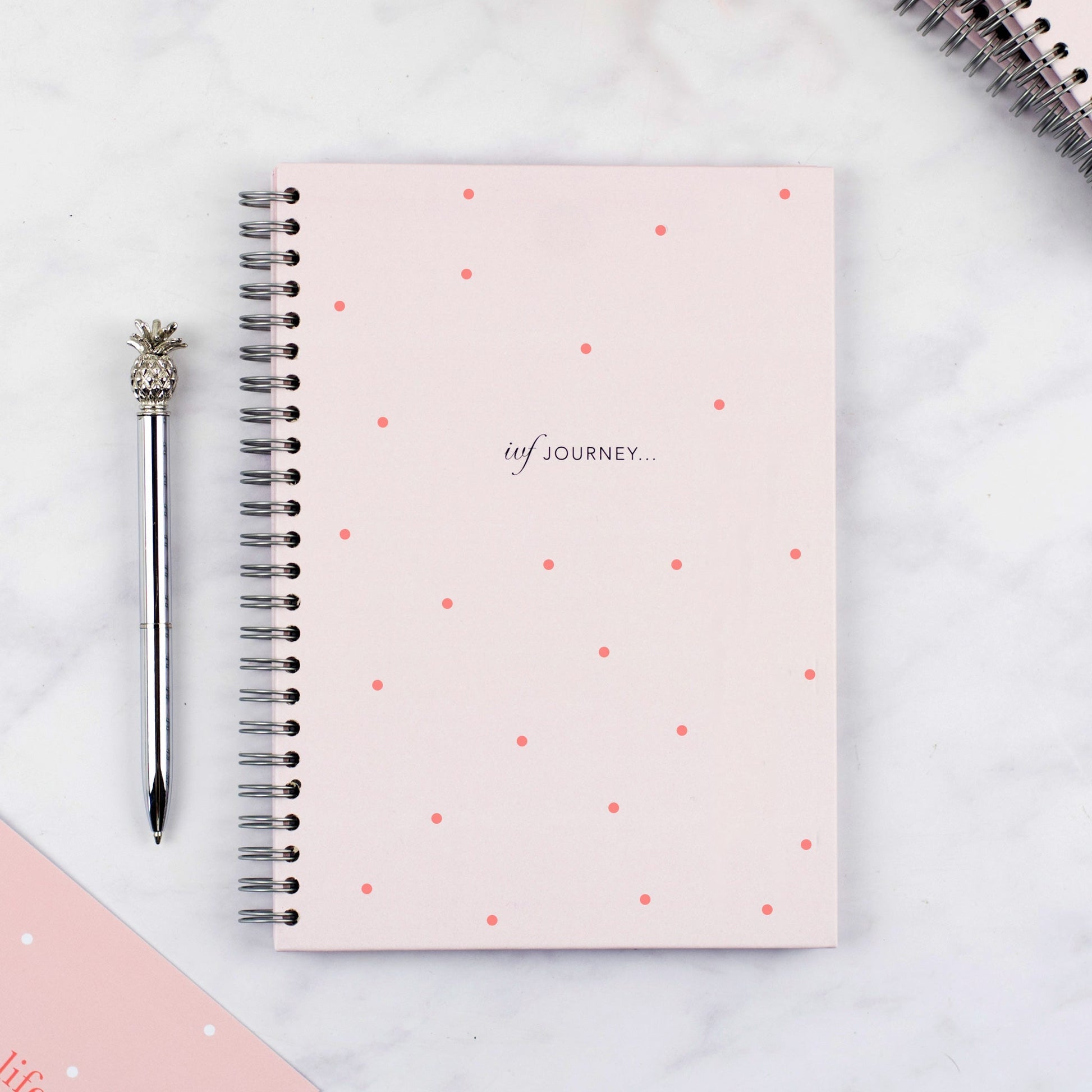 The cover of the pink polka dot IVF Planner with a pineapple pen next to it.