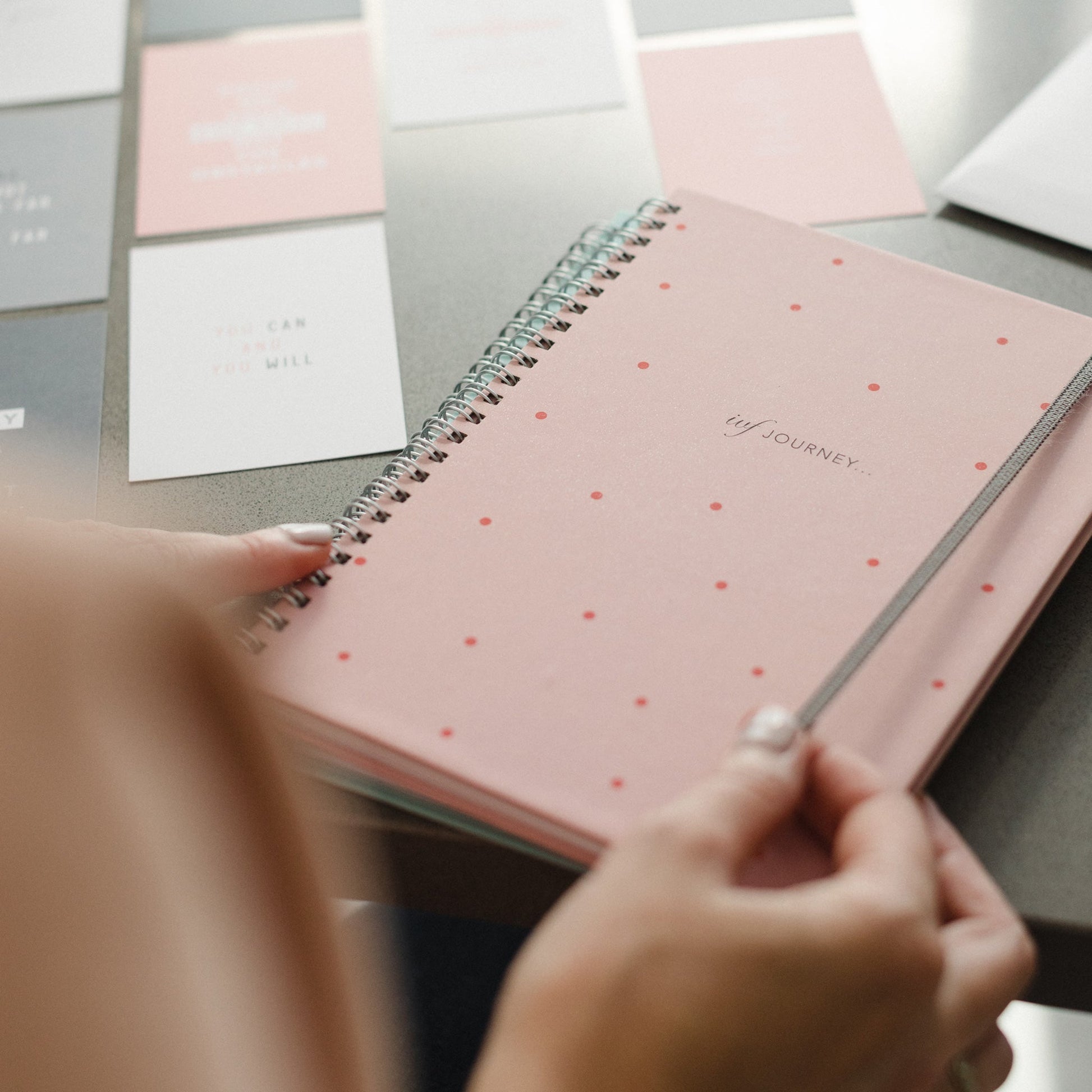 A woman's hand holding the pink polka dot IVF Planner, the two week wait positivity cards are in the background.